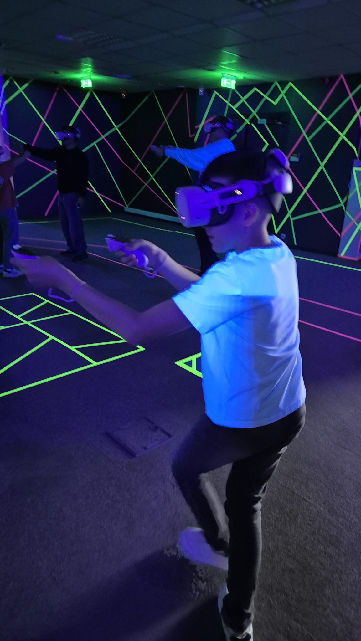 Dalyviai su VR akiniais ir valdikliais žaidžia neoninėje VRspotas arenoje
